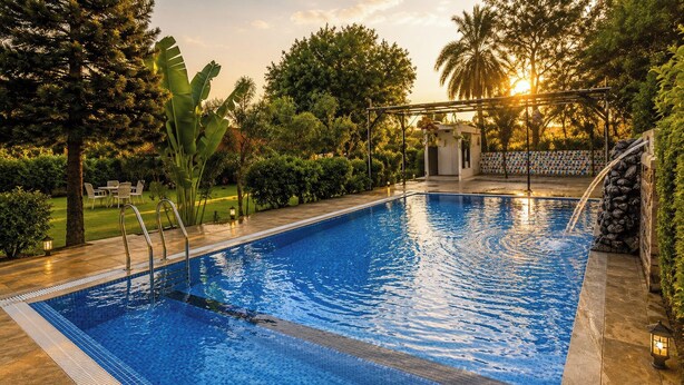 A rectangular swimming pool with clear blue water, surrounded by a tiled deck, and a small pavilion with a decorative roof.