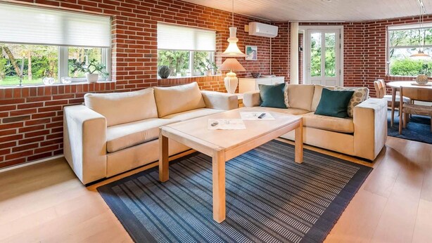 A modern living room with a brick wall, a wooden ceiling, a beige sofa, a coffee table, and a dining area with a table and chairs. - 6 Person Holiday Home in Norre Nebel (Norre Nebel)