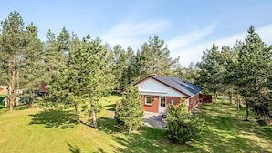 A single-story house with a red brick exterior and a white trim, surrounded by a well-maintained lawn and tall pine trees. - 6 Person Holiday Home in Norre Nebel (Norre Nebel)