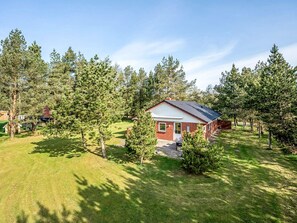 Ein einstöckiges Haus mit roter Backsteinfassade und weißem Zierrahmen, umgeben von einem gepflegten Rasen und hohen Kiefern.