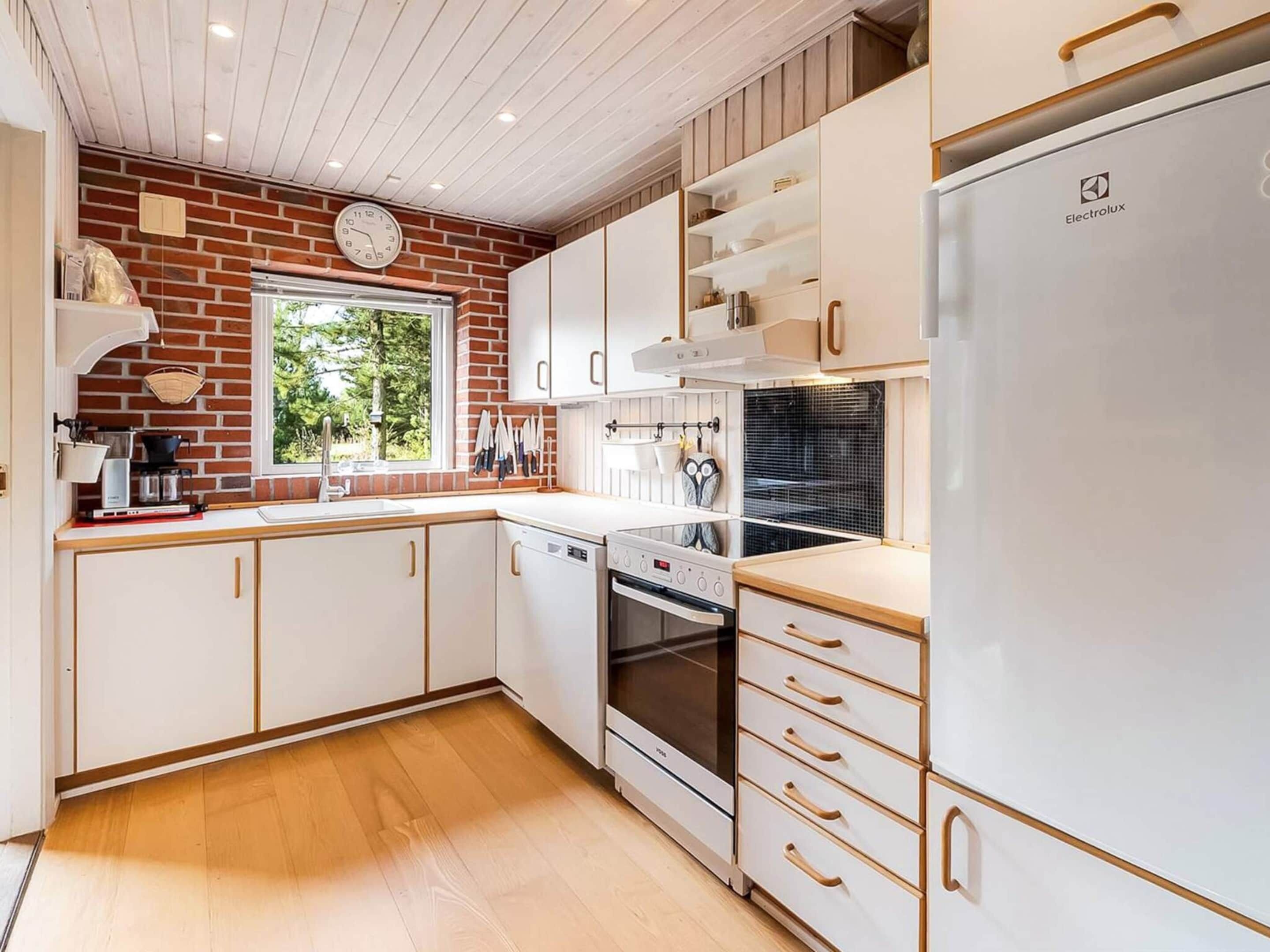 Cocina moderna con armarios blancos, horno integrado, refrigerador y ventana con vista a follaje.