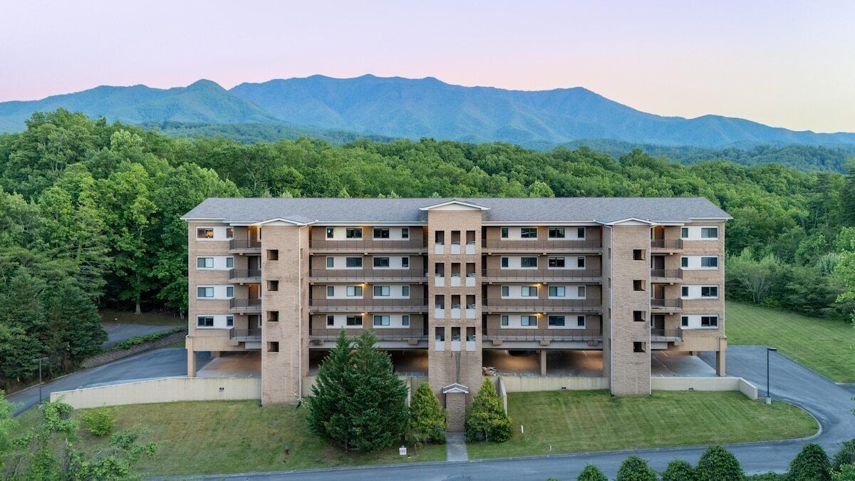 A multi-story hotel with a central entrance, surrounded by lush greenery and mountains in the background.