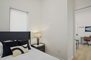 Un dormitorio con cama, mesita de noche, lámpara y vistas a un pasillo que lleva a un baño.