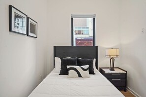 Una cama bien hecha con ropa de cama blanca y almohadas negras y blancas, una mesita de noche con una lámpara y una ventana con vista a un edificio que dice 'PARKING'.