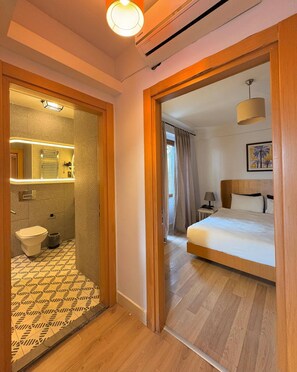 A hotel room with a wooden floor, a bed with white bedding, a bedside table with a lamp, a bathroom with a toilet, a shower, and a mirror with lights. - Taksim Square-1br Rooftop Suite 07 (Istanbul)