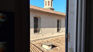 Une vue à travers la balustrade d’un balcon, donnant sur un bâtiment doté d’un toit en tuiles, d’une tour en forme de dôme et d’une fenêtre ornée d’une grille métallique.