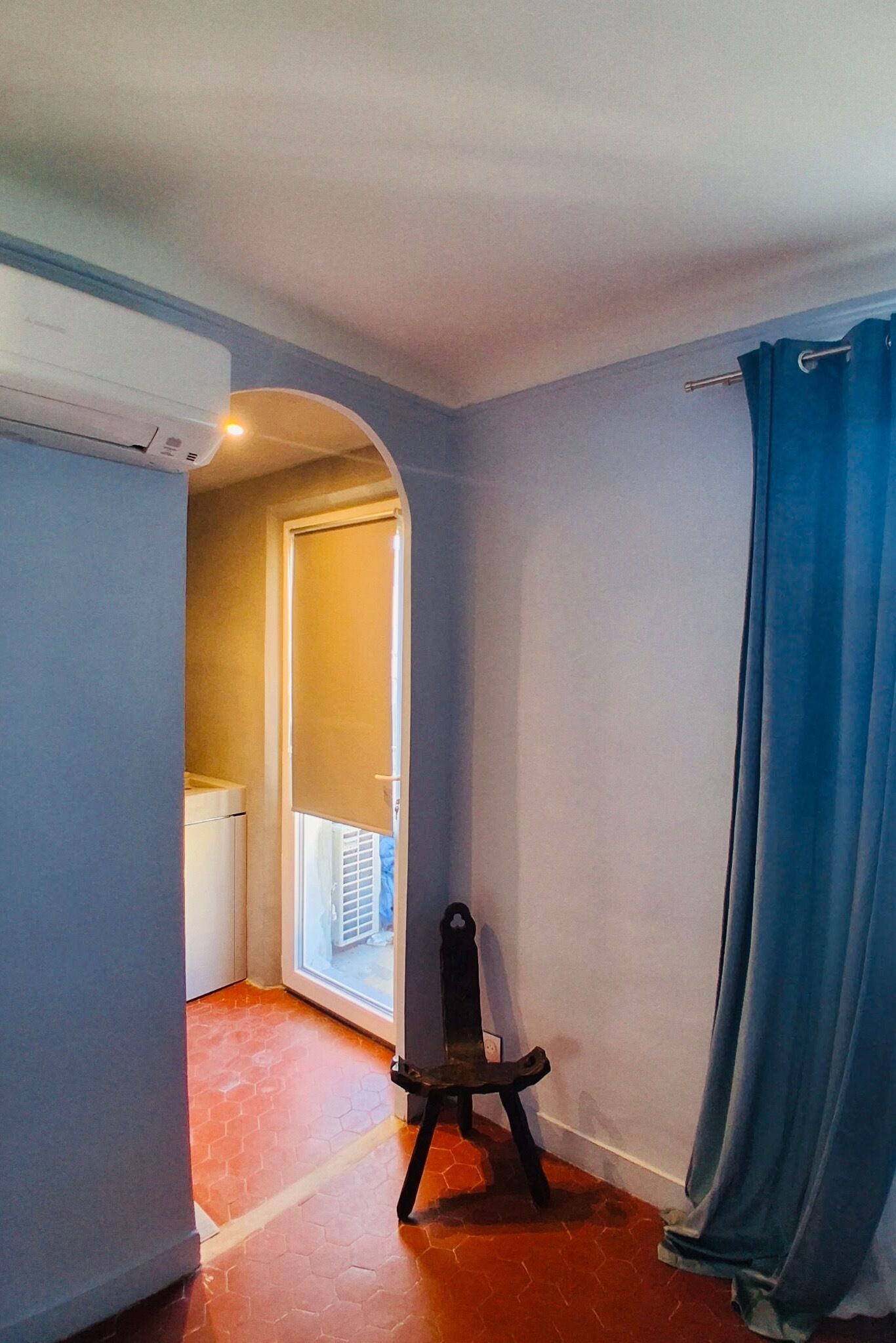 Une pièce avec un sol carrelé, une chaise, un rideau bleu et une porte qui donne sur une autre pièce.