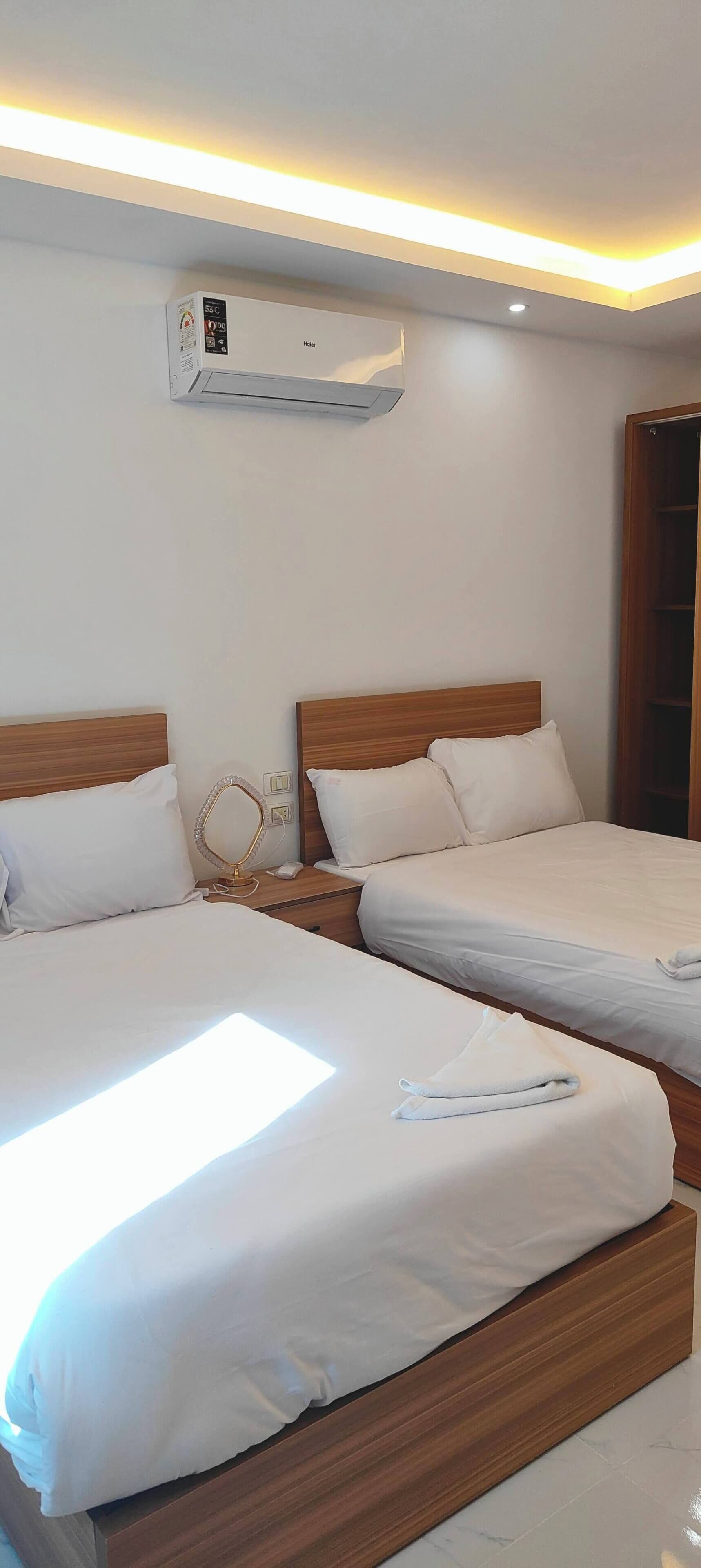 Two beds with wooden headboards, white bedding, a bedside table with a mirror, and an air conditioning unit on the wall.
