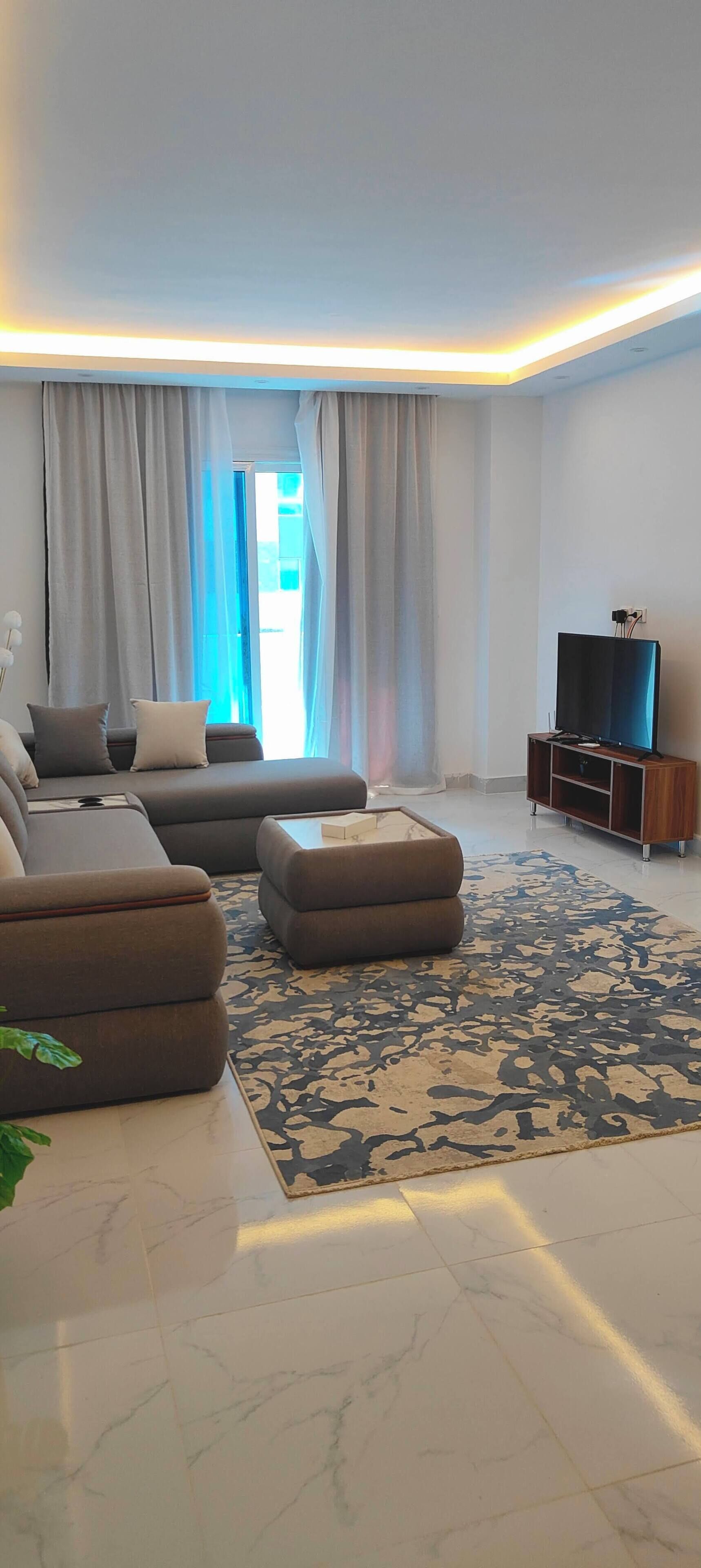 A modern living room with a sectional sofa, ottoman, and a flat-screen TV on a wooden stand.