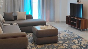 Un salon moderne avec un canapé d’angle, un pouf et un téléviseur à écran plat posé sur un meuble en bois.