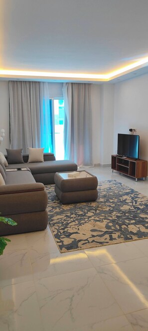 Ein modernes Wohnzimmer mit einem Ecksofa, einem Hocker und einem Flachbildfernseher auf einem Holzboden.