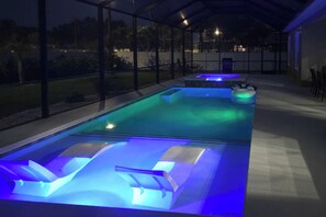 A swimming pool with built-in seating and blue lighting, surrounded by a covered area with glass walls. - Private Outdoor Oasis! Home 12 Mi to Flagler Beach (Palm Coast)