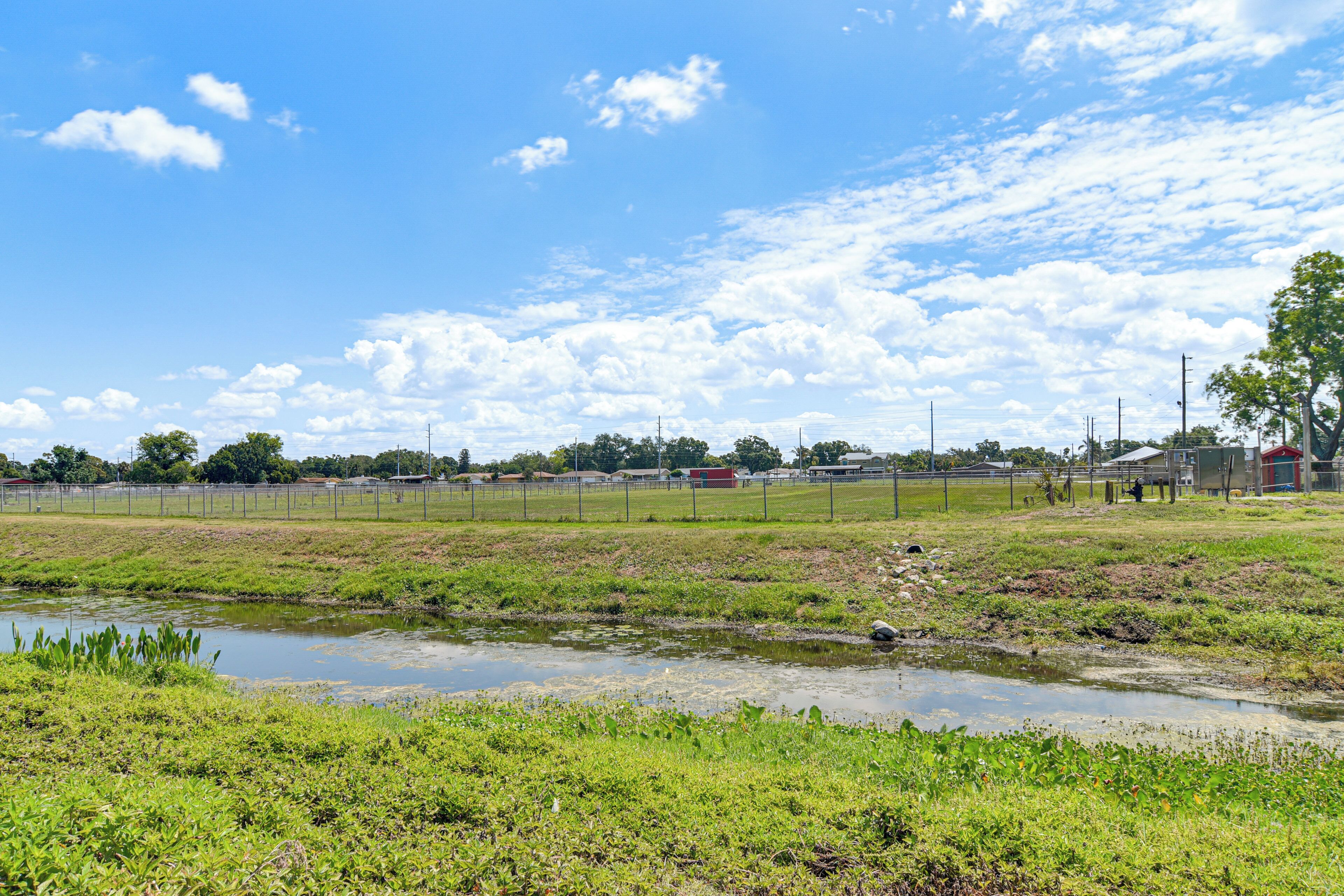 A rural landscape with a small pond, grassy fields, and distant buildings.