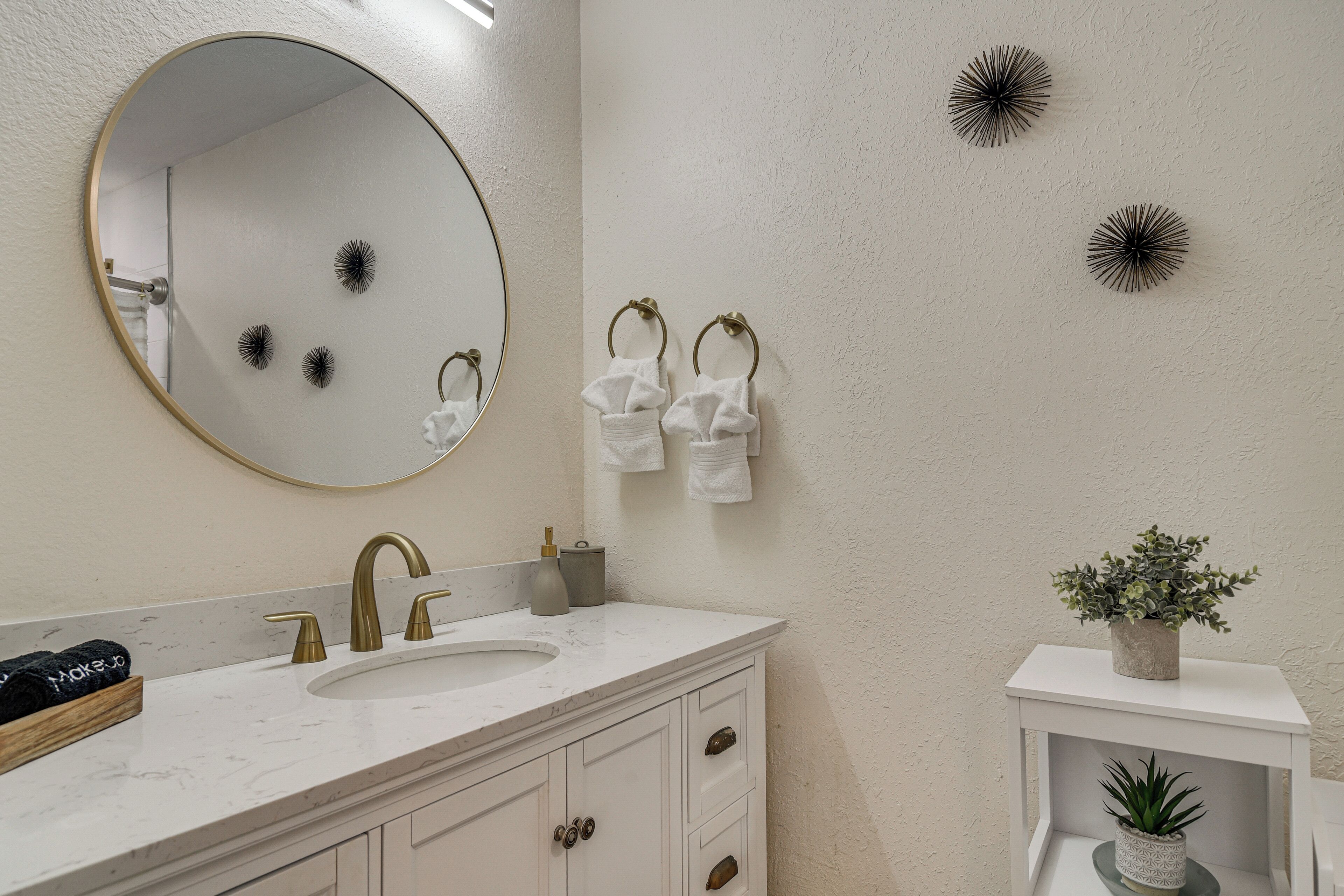 A bathroom with a round mirror, a sink with a gold faucet, white towels, decorative wall art, and a small plant on a shelf.