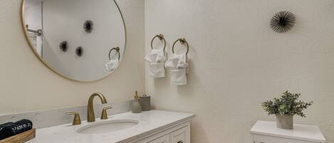 A bathroom with a round mirror, a sink with a gold faucet, white towels, decorative wall art, and a small plant on a shelf.