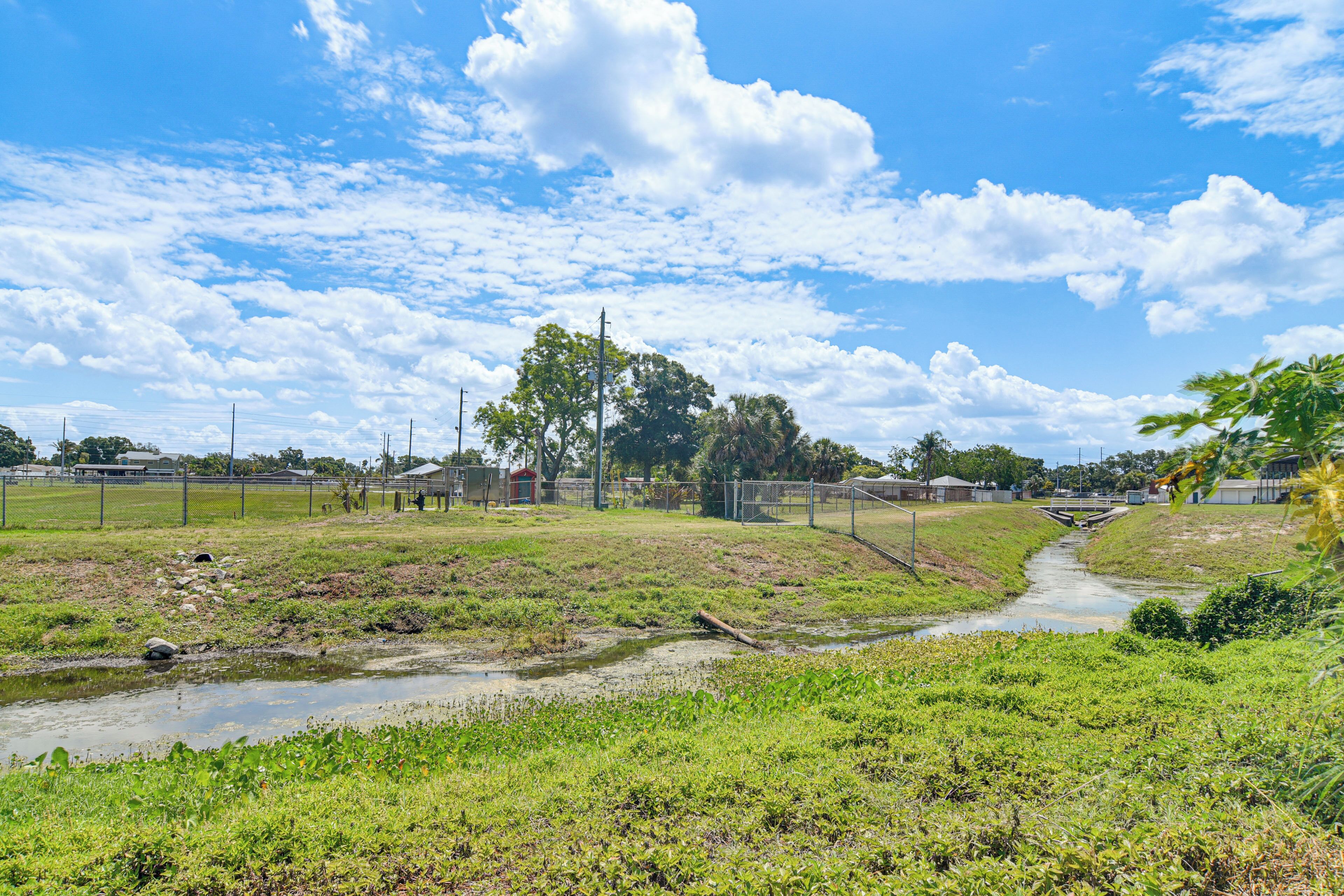 Un paysage rural avec un petit canal d’irrigation, des champs couverts d’herbe et un ensemble de bâtiments situé au loin.