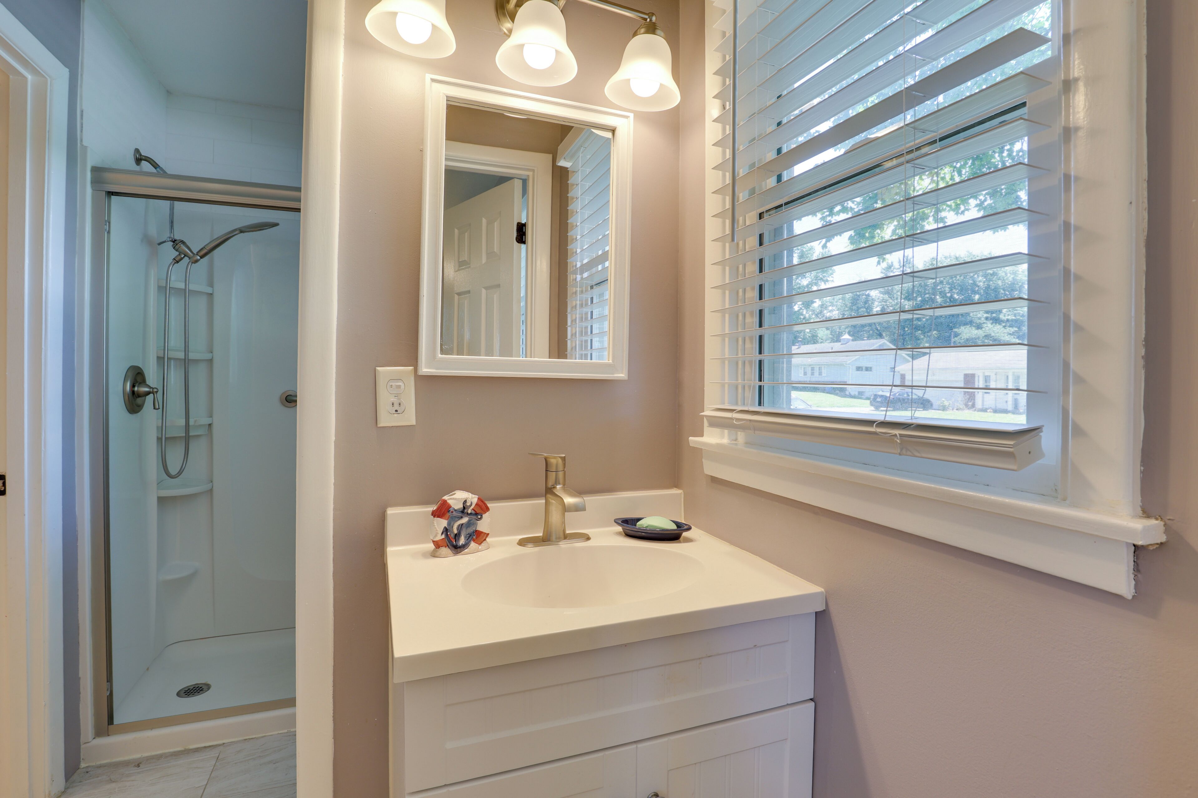 A bathroom with a shower, a sink with a mirror, and a window with blinds.