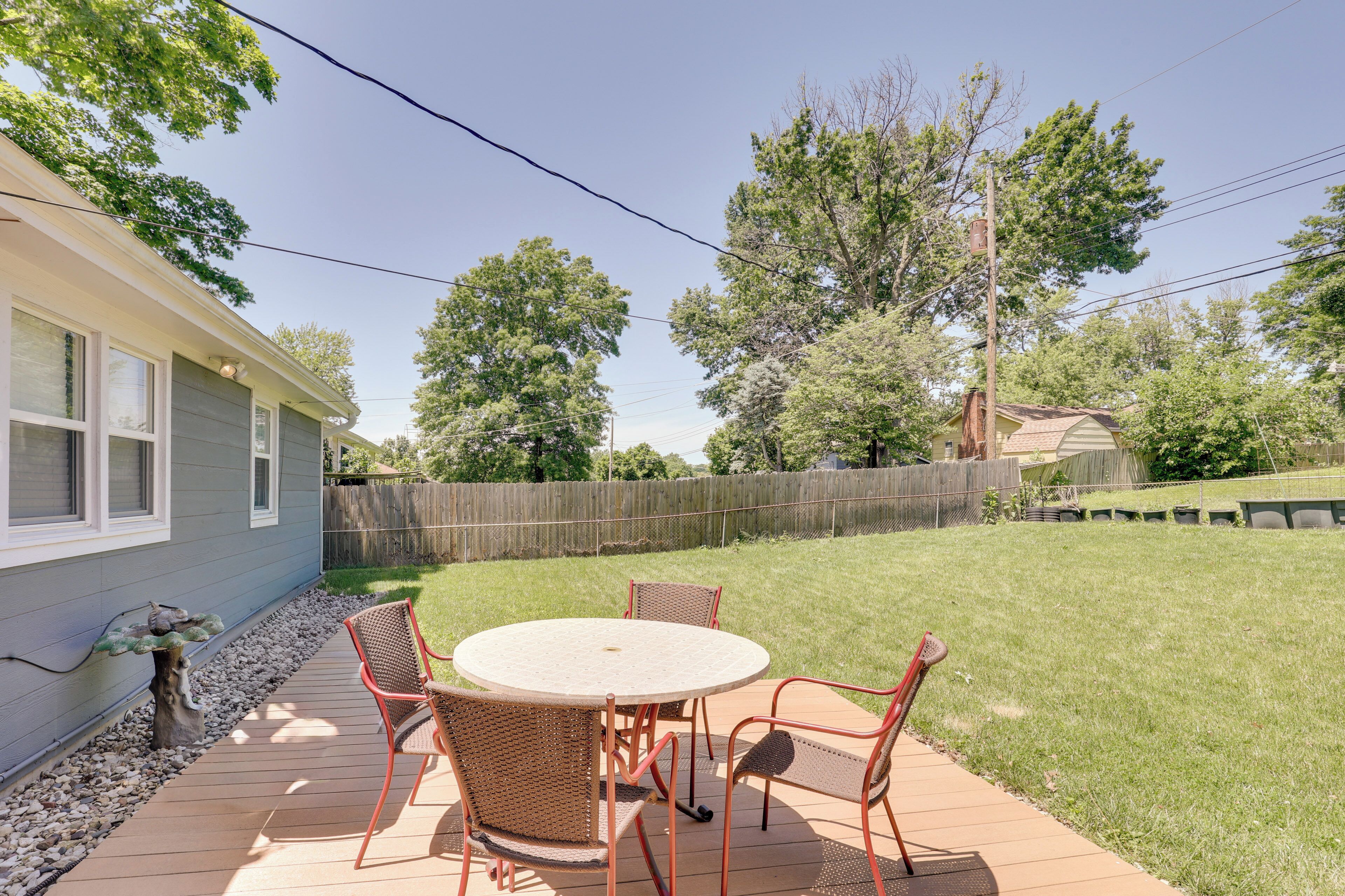 A patio with a round table and chairs, a wooden deck, and a backyard with a wooden fence and trees.