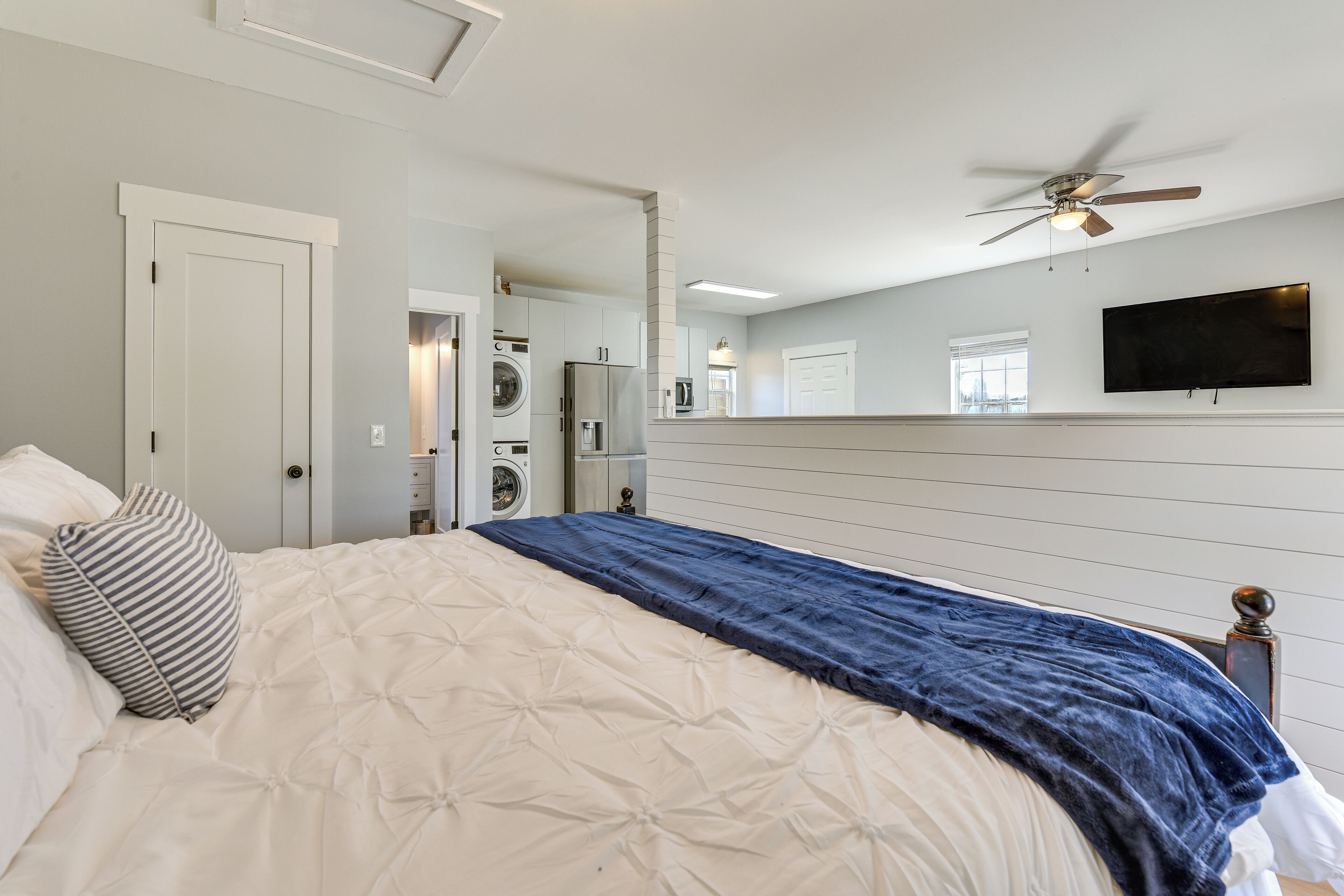 Ein Schlafzimmer mit einem Bett, einem Deckenventilator, einem an der Wand befestigten Fernseher und einem Waschbereich, der durch eine geöffnete Tür sichtbar ist.