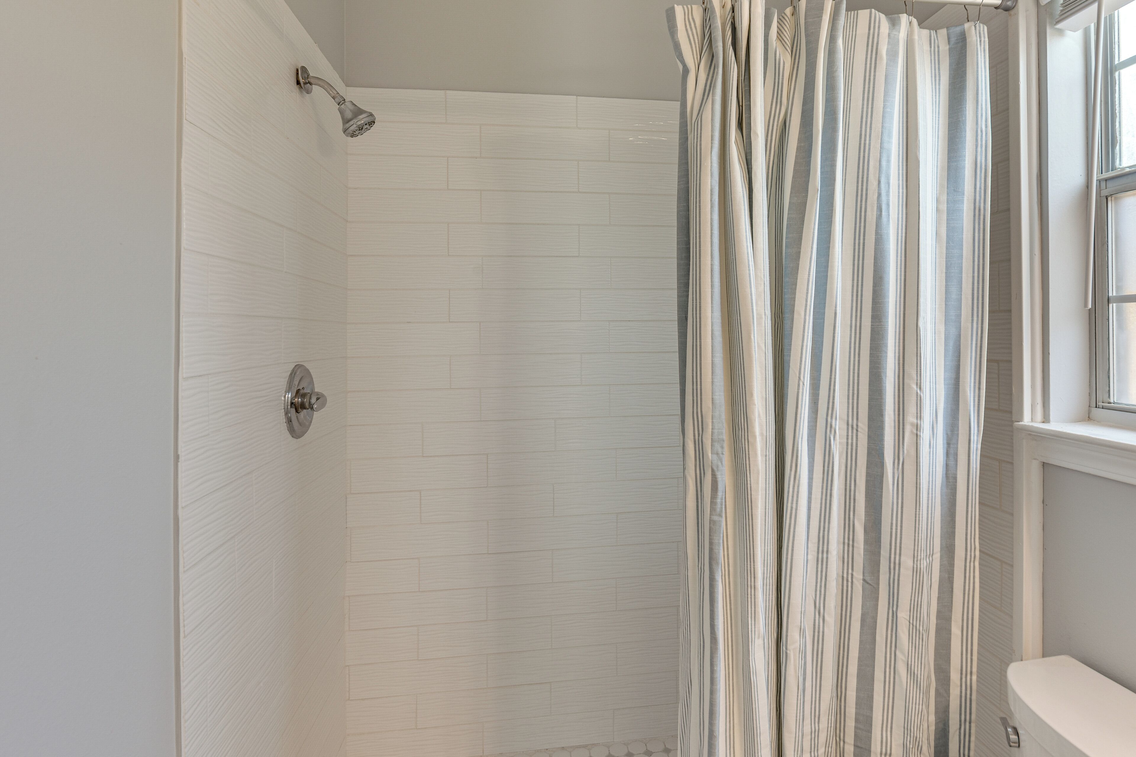 A bathroom with white tiled walls, a shower with a striped curtain, and a window with blinds.
