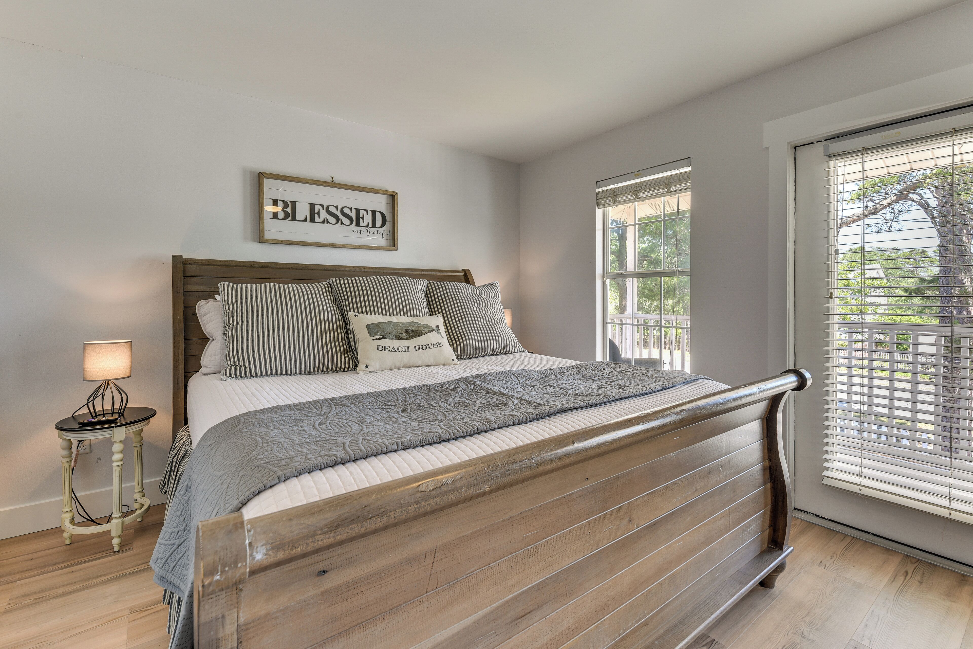 Une chambre avec un lit en bois, une table de chevet avec une lampe, un cadre décoratif accroché au mur et une fenêtre avec des stores.