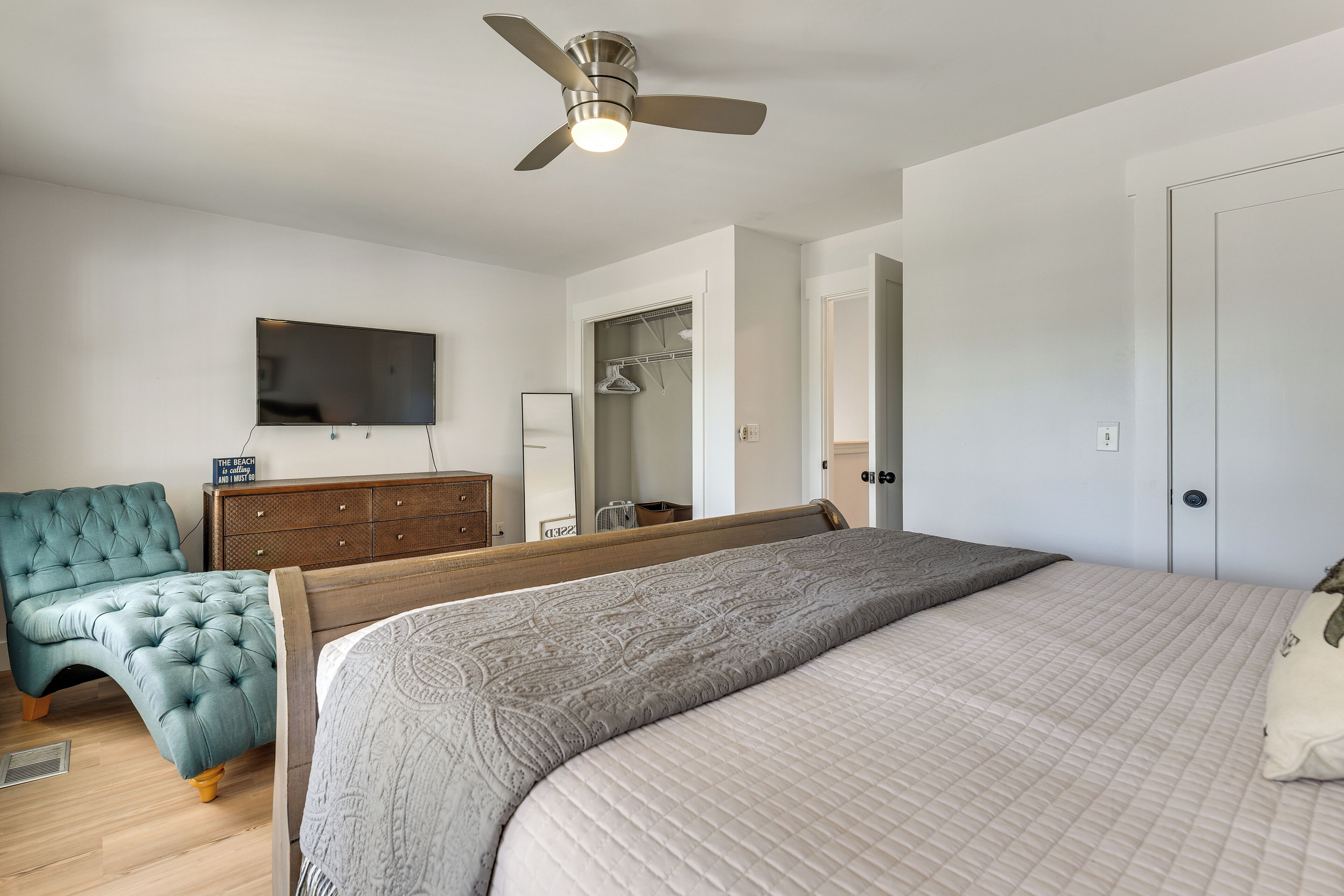 Une chambre à coucher comprenant un lit, un fauteuil capitonné, une commode, un téléviseur, un ventilateur de plafond et un dressing.