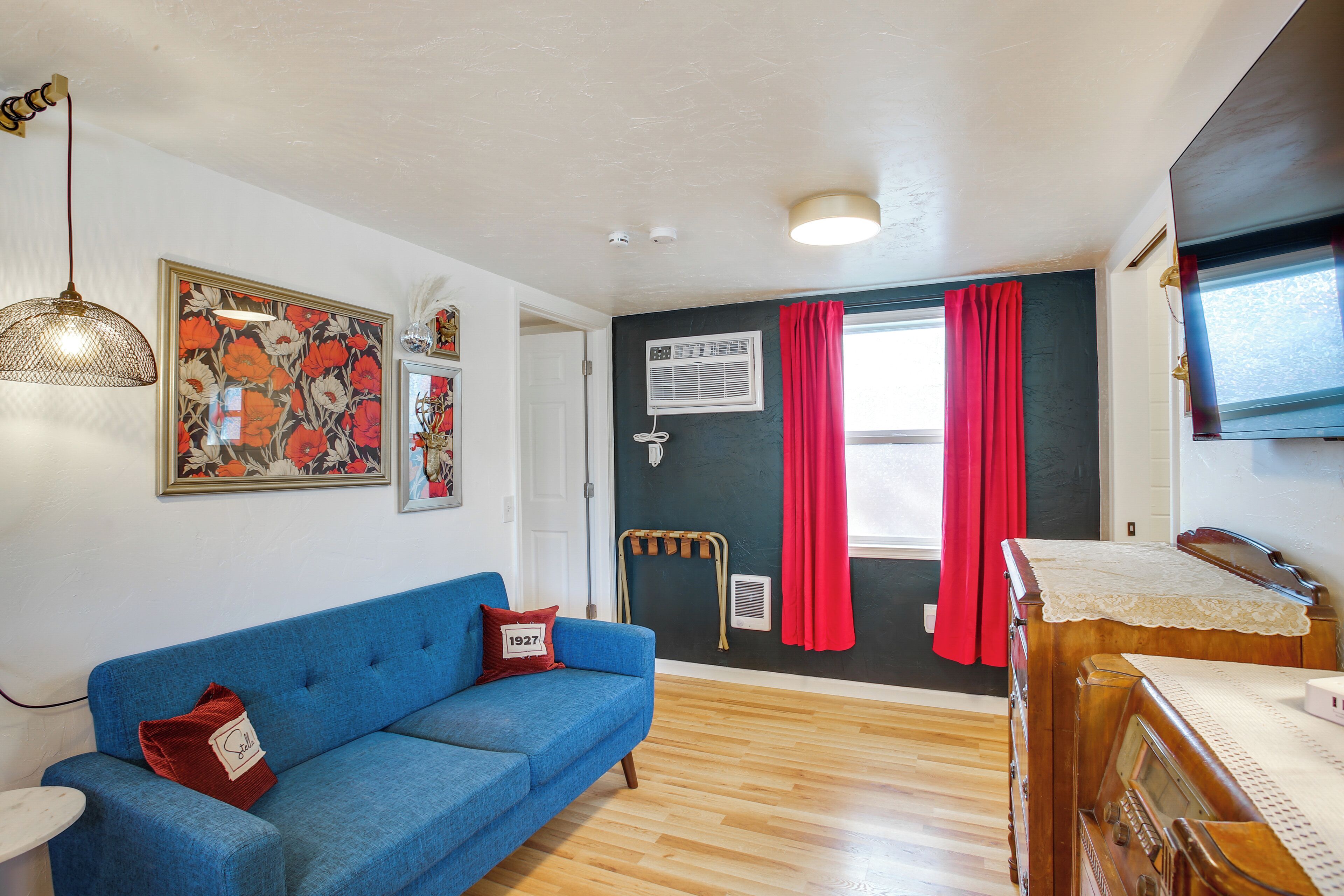 Una habitación con un sofá azul, un aparador de madera, una lámpara de techo y una ventana con cortinas rojas.