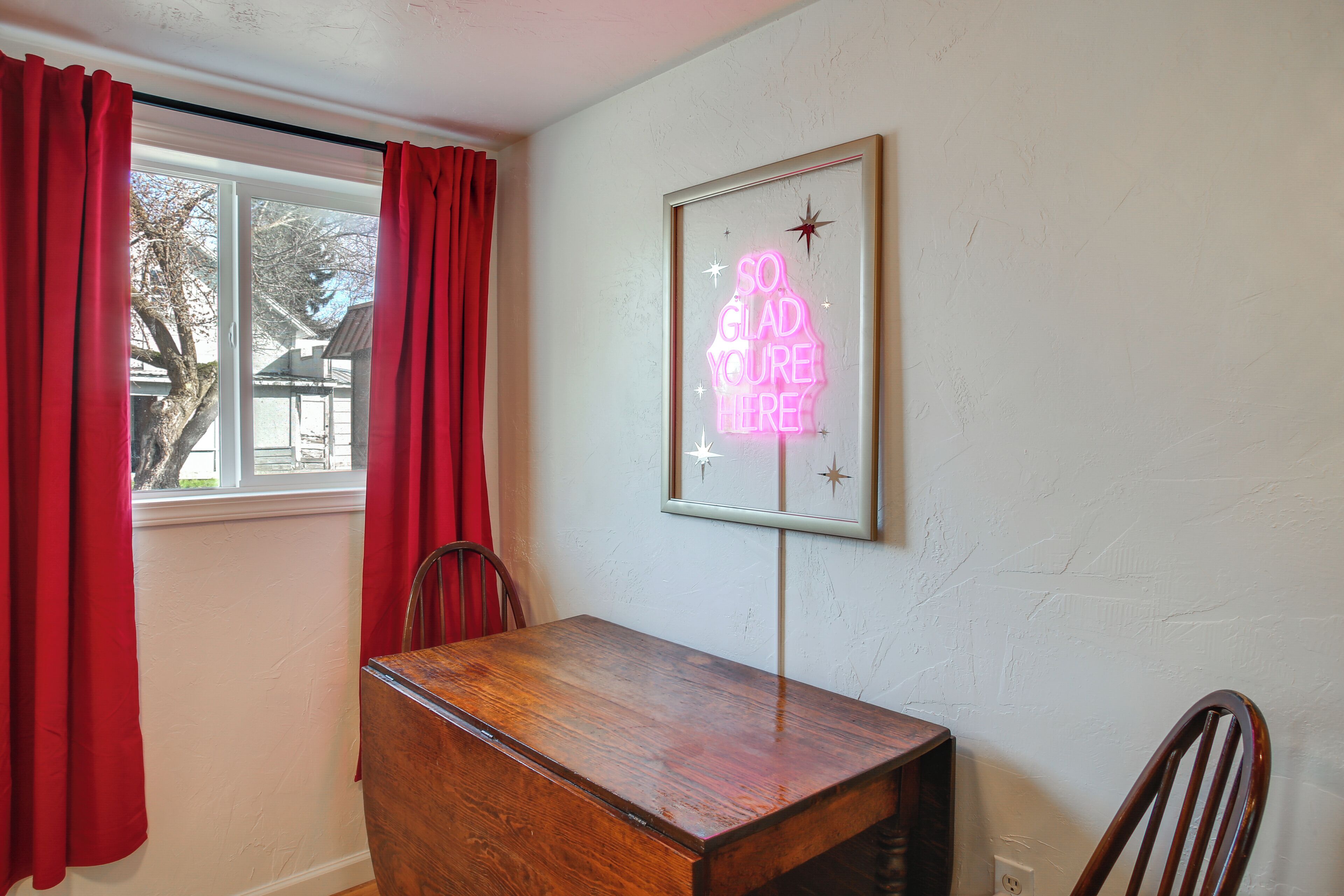 A room with a wooden table, red curtains, a framed neon sign, and a chair.