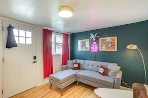 Una habitación con un sofá seccional gris, una puerta blanca, una ventana con cortinas rojas, una lámpara de pared y un letrero de neón.