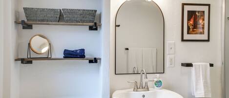 Une salle de bain comprenant des toilettes, un lavabo, un miroir, des étagères avec des paniers et un tableau encadré au mur.