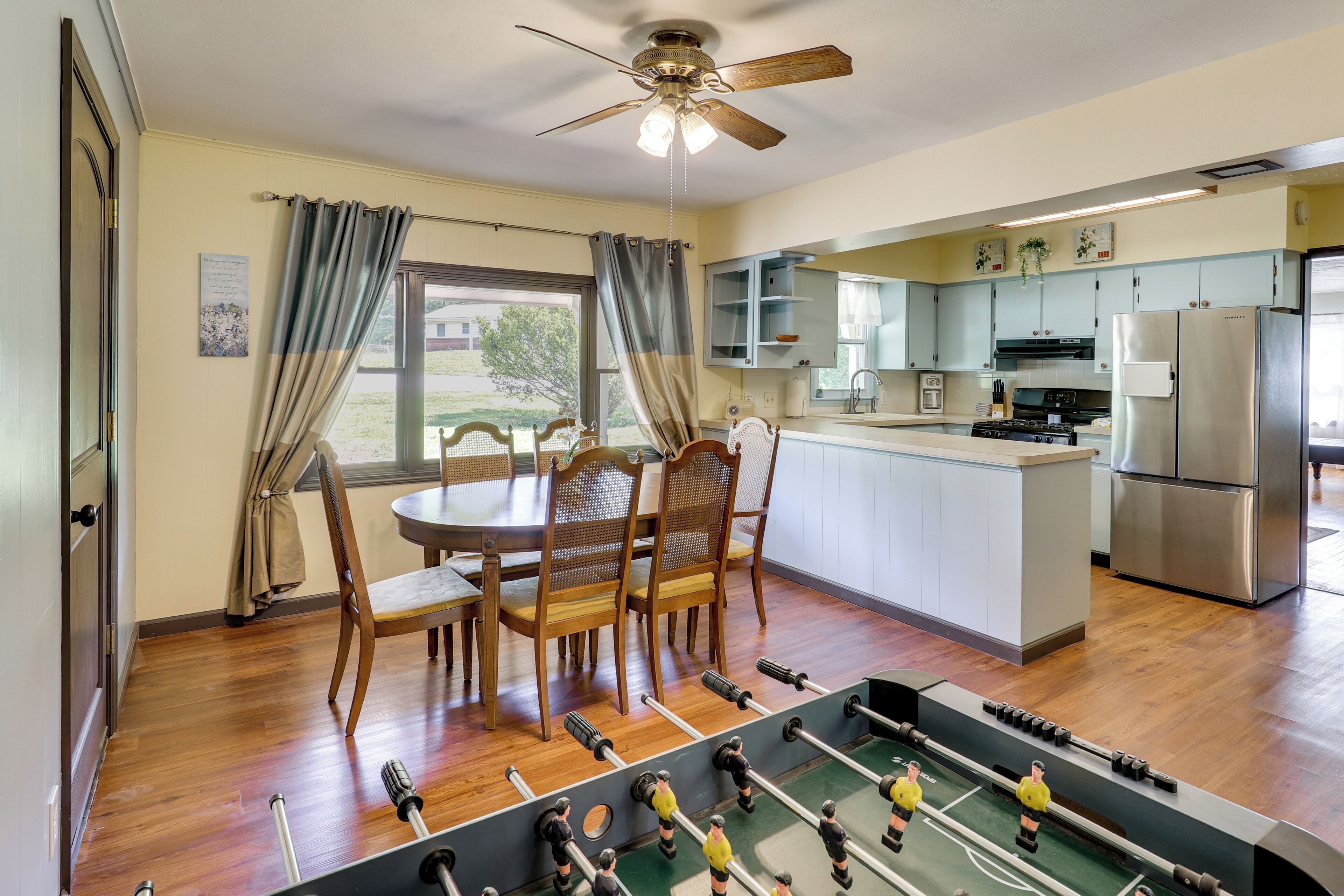 A room with a foosball table, wooden chairs, a dining table, a ceiling fan, and a kitchen area with a refrigerator and cabinets.