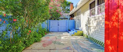 Un patio trasero residencial con una verja azul, un cobertizo rojo y un patio de hormigón con obras de arte coloridas.