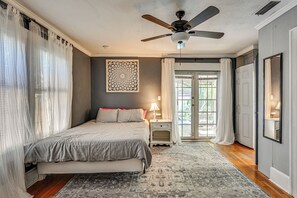 Ein Schlafzimmer mit Bett, Deckenventilator, Nachttisch, Lampe, einem gemusterten Teppich und einer Tür, die auf einen Balkon führt.