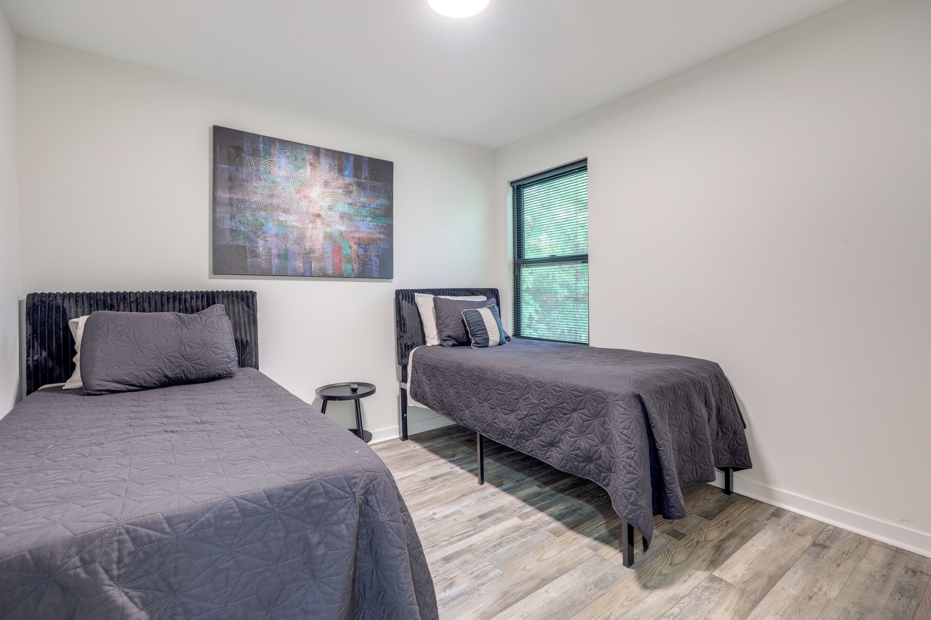 A bedroom with two beds, a small round table, a large abstract painting, and a window with blinds.