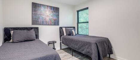 A bedroom with two beds, a small round table, a large abstract painting, and a window with blinds.