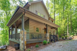 A two-story log cabin with a porch, stone foundation, and a forested backdrop.