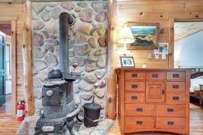 A rustic cabin interior with a stone wall, a wood-burning stove, a wooden dresser, and a painting on the wall.