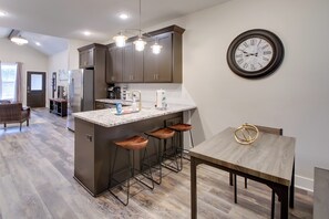 A modern kitchen with dark cabinets, a central island with stools, a wall-mounted clock, and a dining area with a wooden table. - 2-unit Hot Springs Retreat Near Bathhouse Row (Hot Springs)