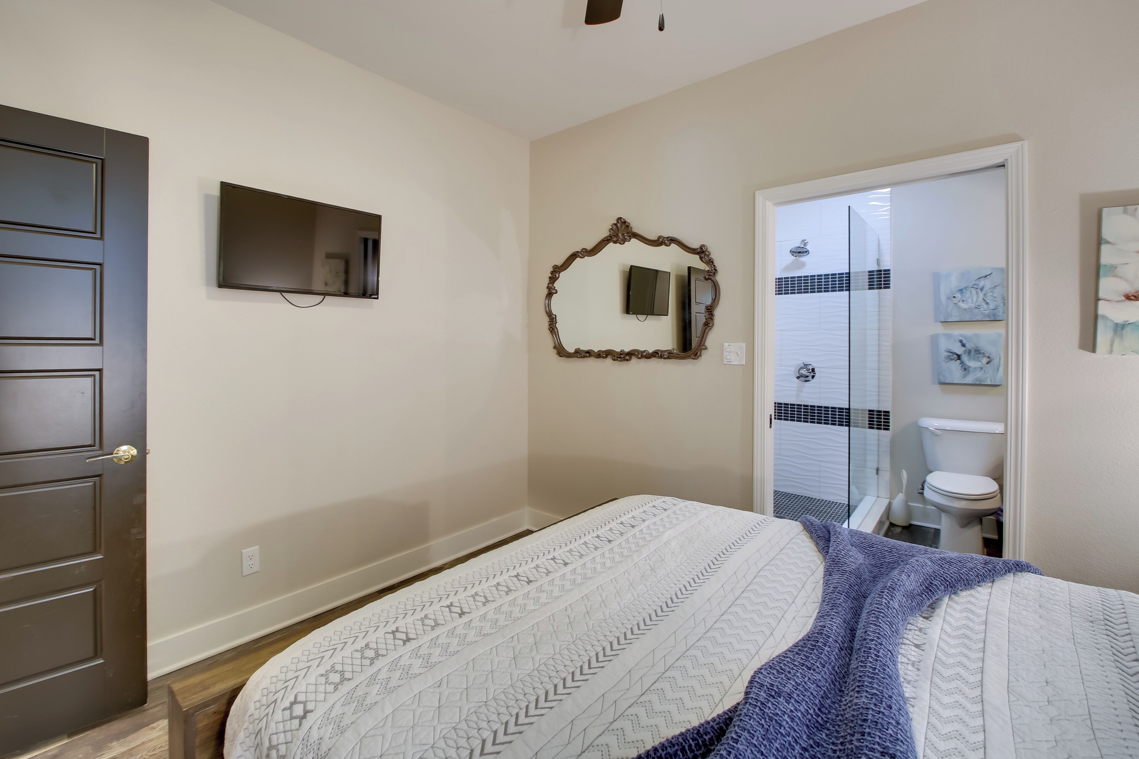Une chambre à coucher avec un lit, une télévision, un miroir et une salle de bain visible à travers une porte ouverte.