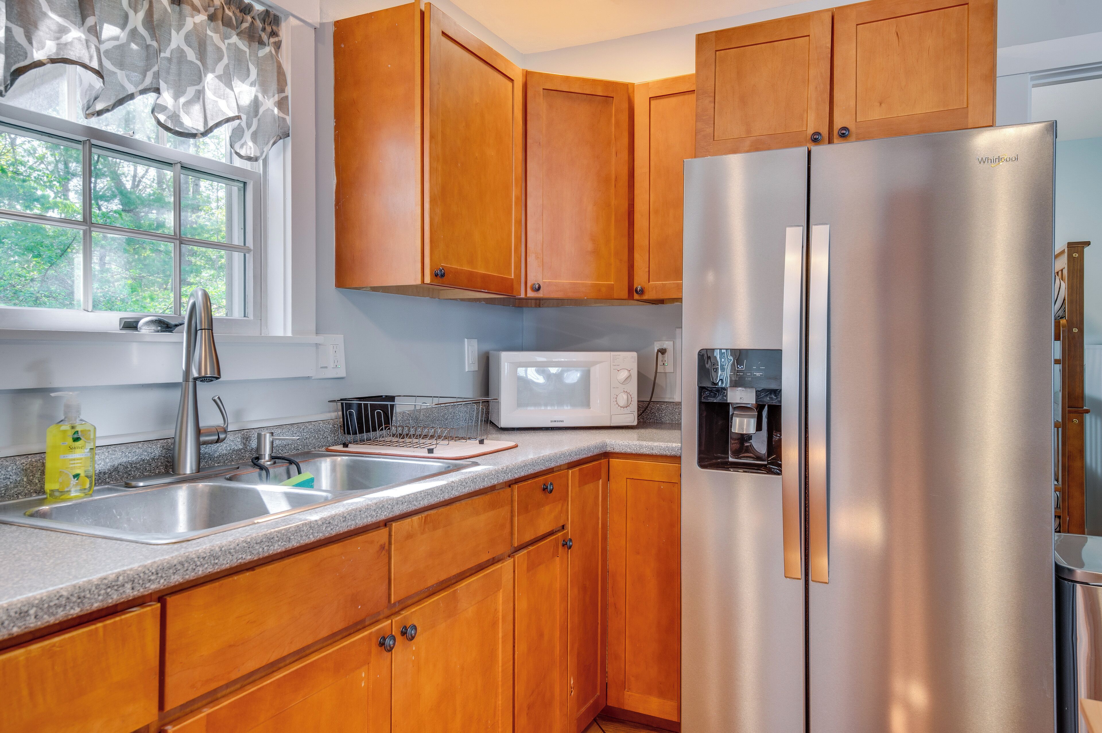 A kitchen with wooden cabinets, a stainless steel refrigerator, a microwave, and a window with a view of trees.