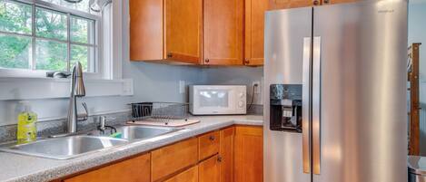 A kitchen with wooden cabinets, a stainless steel refrigerator, a microwave, and a window with a view of trees.