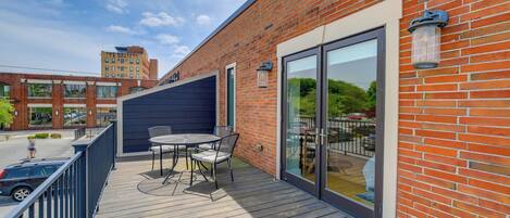 Una terraza de madera con mobiliario exterior, una puerta de cristal y un edificio de ladrillo al fondo.