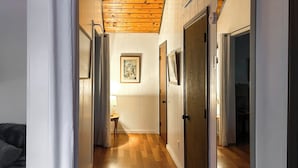 A hallway with wooden flooring, white walls, and a ceiling with exposed beams. There are doors on both sides, a framed picture on the wall, and a small table with a lamp. - Tranquil Mtn Guest Suite Outside Morganton (Connellys Springs)