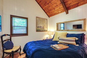 Ein Schlafzimmer mit Holzbalkendecke, einem Bett mit blauer Bettwäsche, einem Sessel mit Kissen, einem Nachttisch mit Lampe und einem Bild an der Wand.