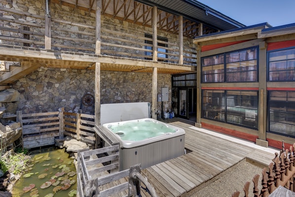 A hot tub on a wooden deck, stone walls, and a building with large windows. - Hike, Soak + Play: Rustic Maysville Escape! (Maysville)