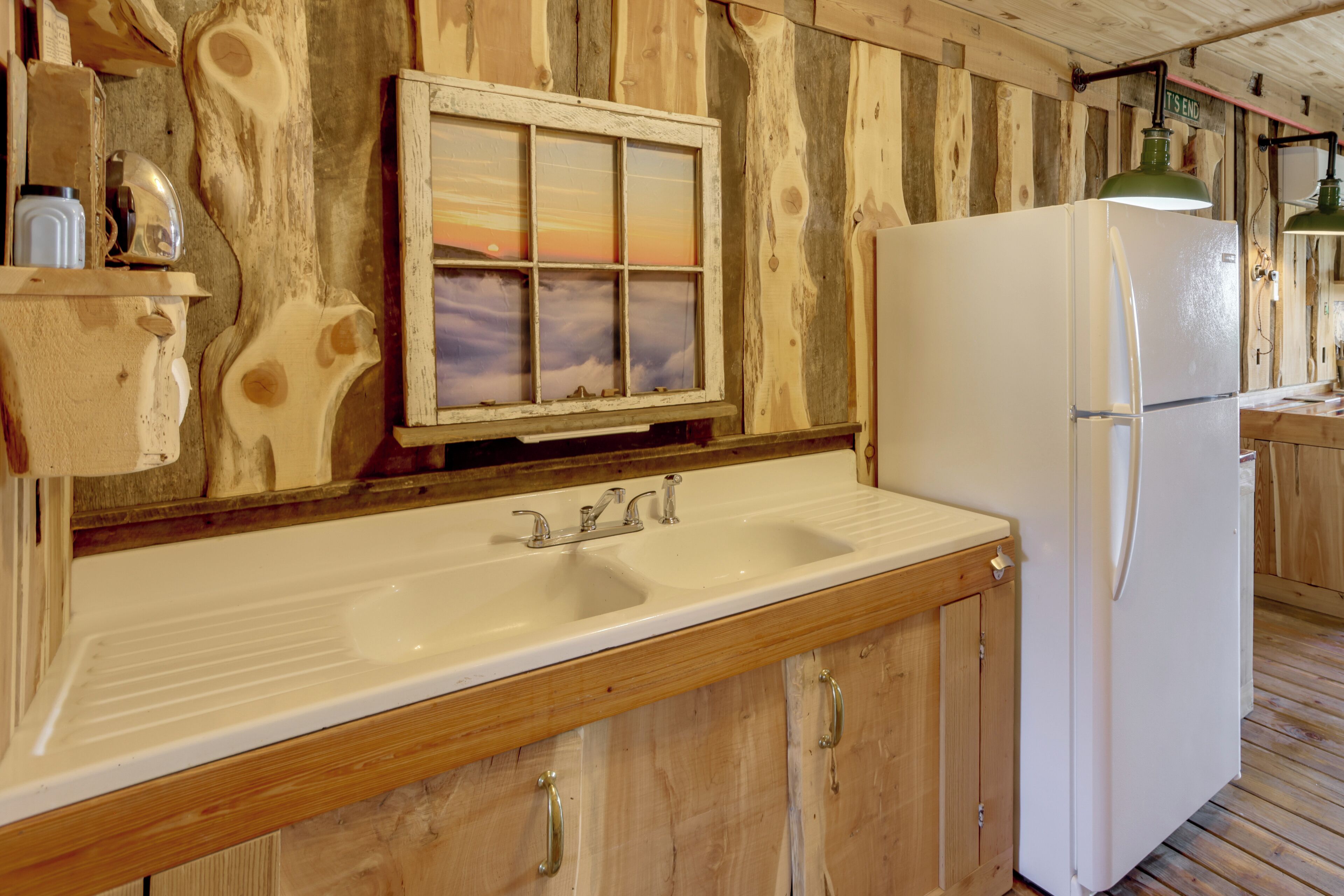 A wooden interior with a white sink, a window with a sunset view, and a white refrigerator.