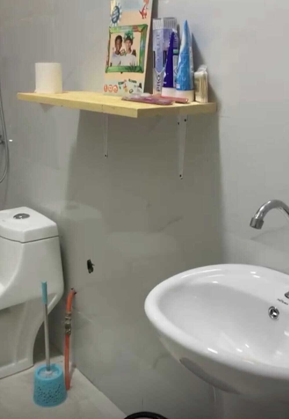 Un baño con lavamanos blanco, inodoro, ducha y repisa con artículos de aseo.