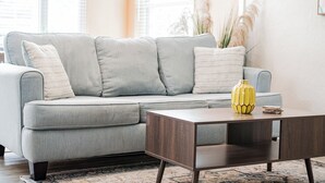 Ein hellgraues Ecksofa mit vielen Kissen, ein Couchtisch aus Holz mit einer gelben Vase und ein gemustertes Teppich auf Holzdielen.