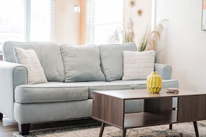 Ein hellgraues Ecksofa mit vielen Kissen, ein Couchtisch aus Holz mit einer gelben Vase und ein gemustertes Teppich auf Holzdielen.