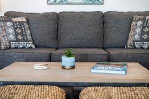 Ein Wohnzimmer mit grauem Sofa, gemusterten Kissen, einem Holztisch, einer kleinen Pflanze und einem Buch.